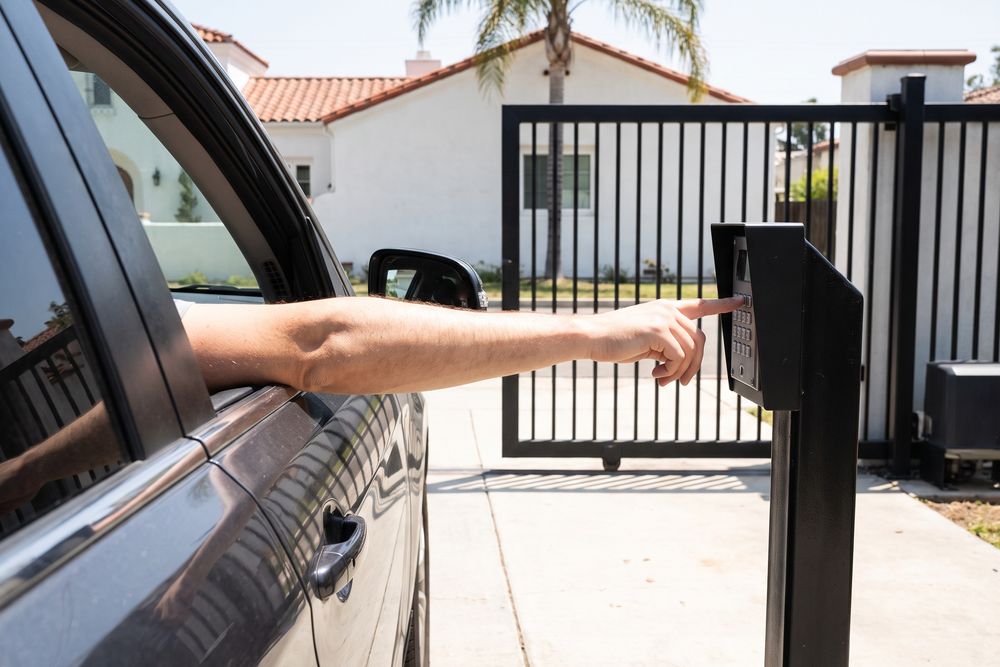 A driver reaches out of the car window to press the keypad at a black steel driveway gate